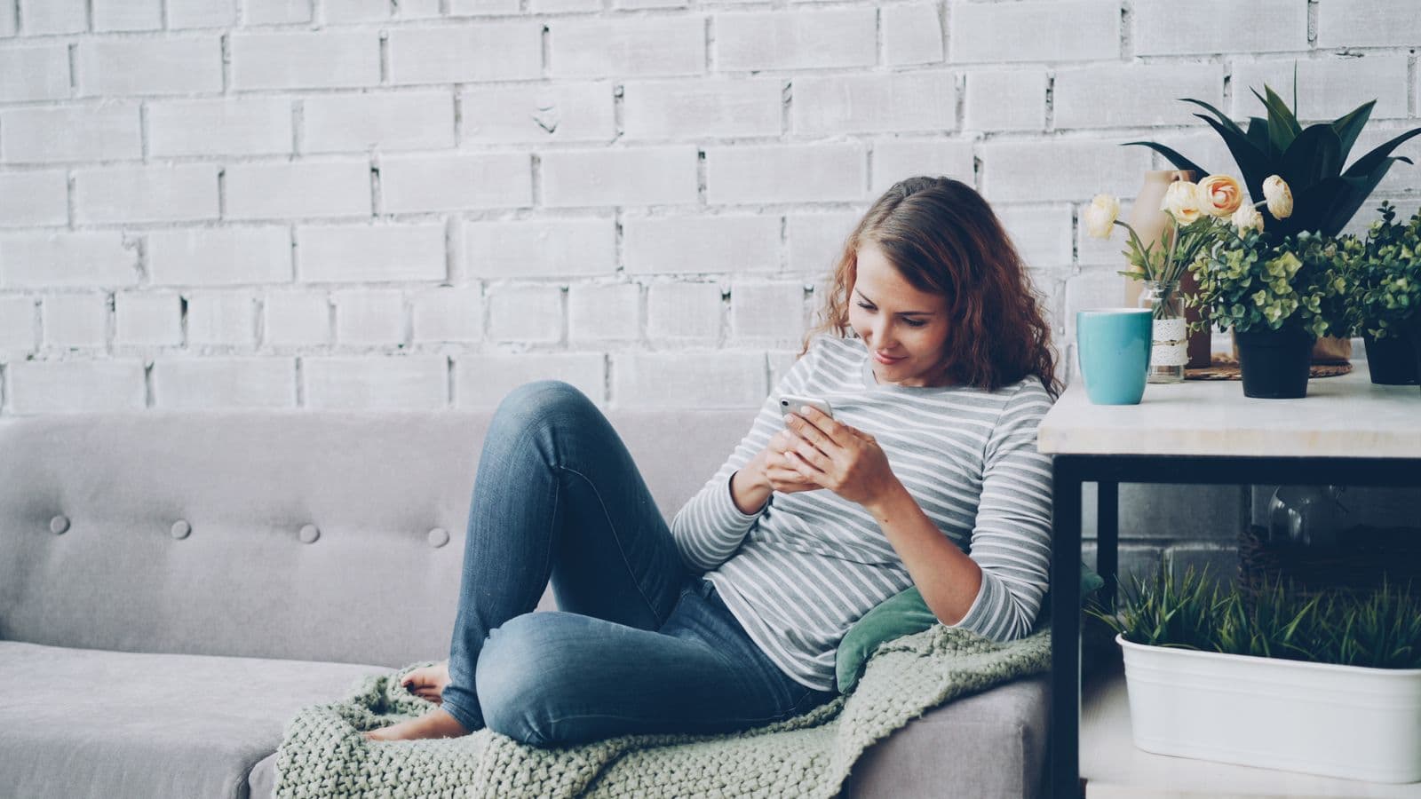 Eine Frau sitzt entspannt auf dem Sofa und schaut lächelnd auf ihr Handy